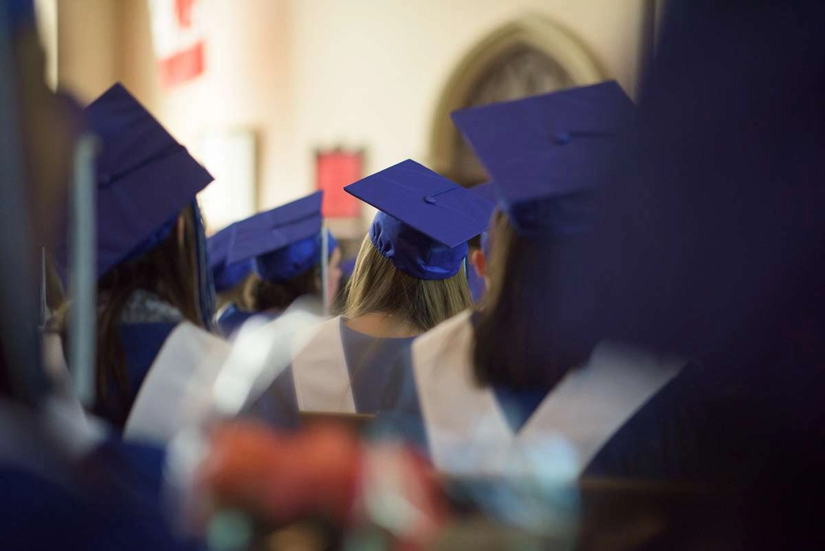 Students donning caps and gowns, sit waiting for their names to be called during the graduation ceremony. The photograph is captured at eye-level through rows of seated students, offering an interesting perspective and creating a sense of being imme…