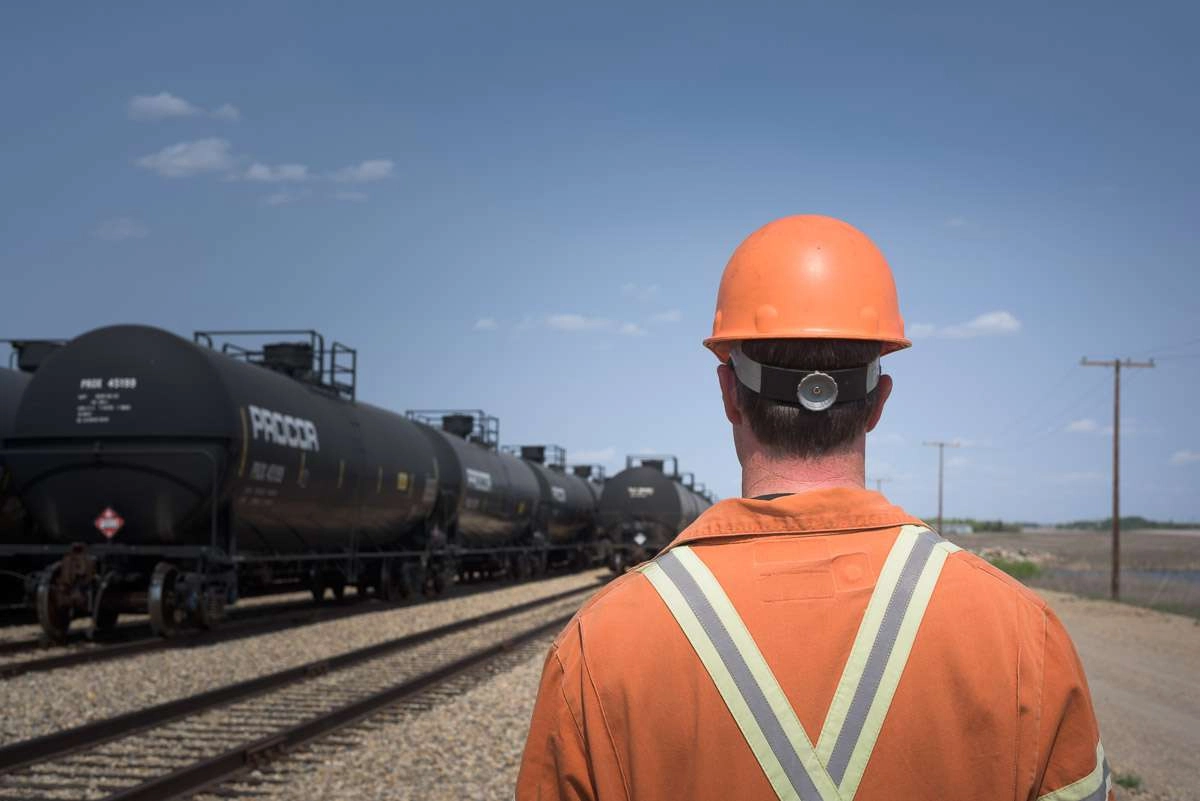 A crew member in an orange jumpsuit and hard hat looks towards the tracks and trains. This photograph captures the employee’s view from over his shoulder as he walks along the tracks.