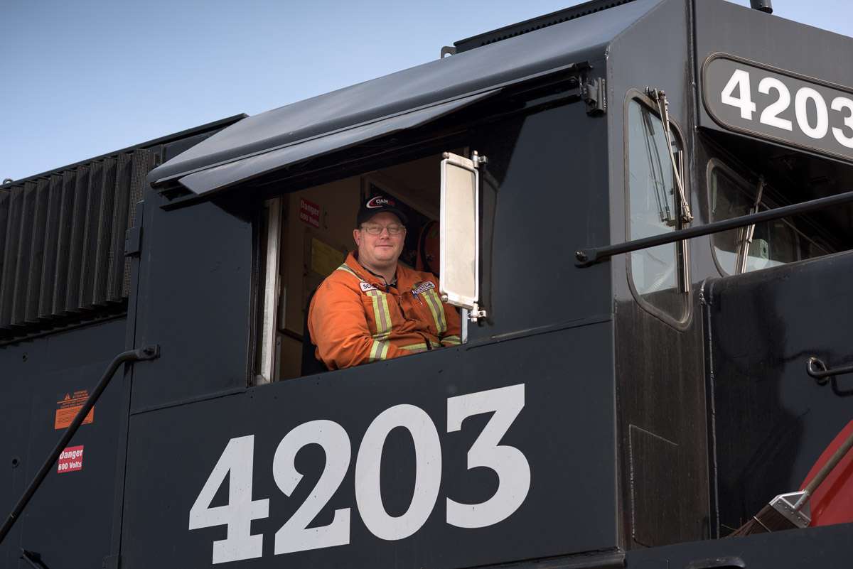 A crew member in an orange jumpsuit sits in the first car of a cando train. The photograph is taken at a low angle, to include some of the exterior of the train, and provide a sense its height.