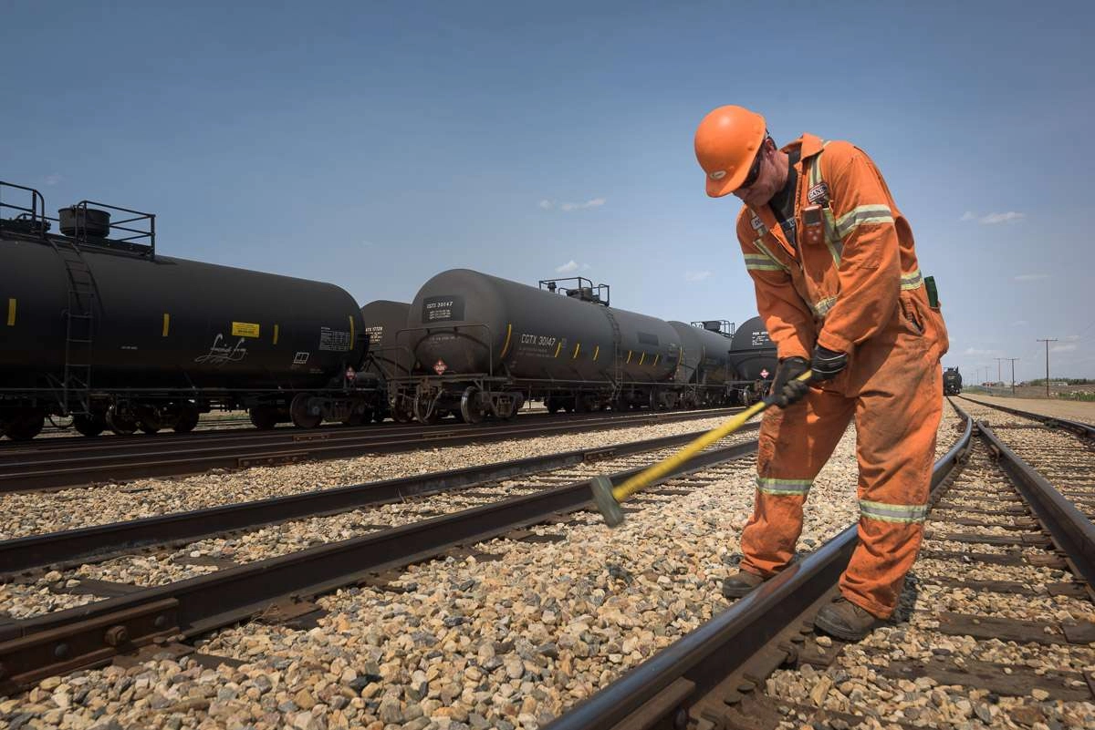 An employee in an orange jumpsuit and hard hat works on the tracks. The rails create leading lines in the photograph, guiding the eye to the employee hard at work. Tank cars line the tracks behind him.