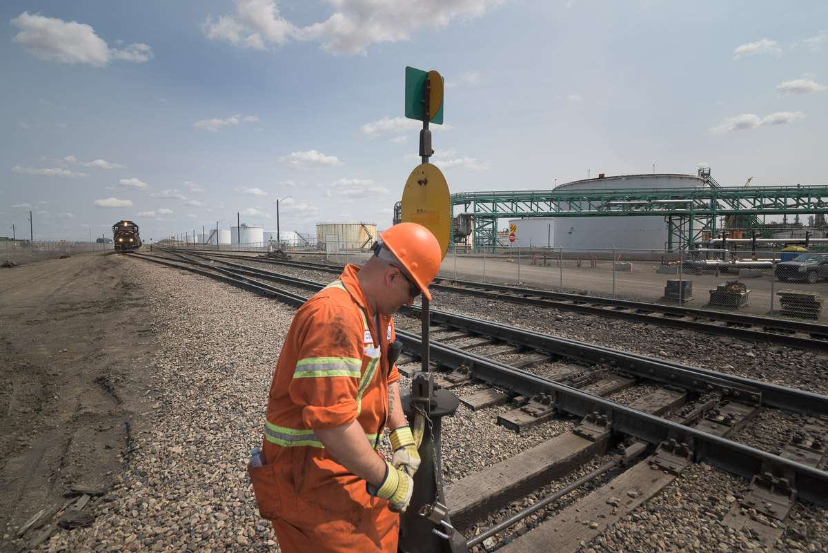 A train approaches in the distance, while an employee works beside the track. This image depicts the vastness of the yard, with an assortment of equipment and facilities visible in the background.