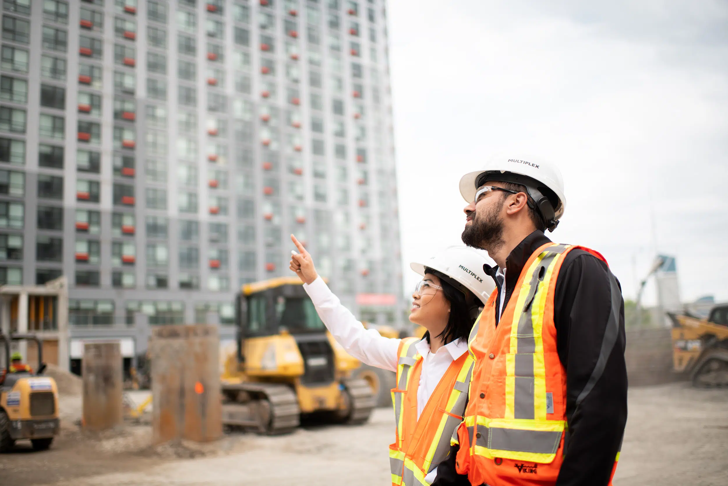 A female and male worker in orange vests discuss a construction project.