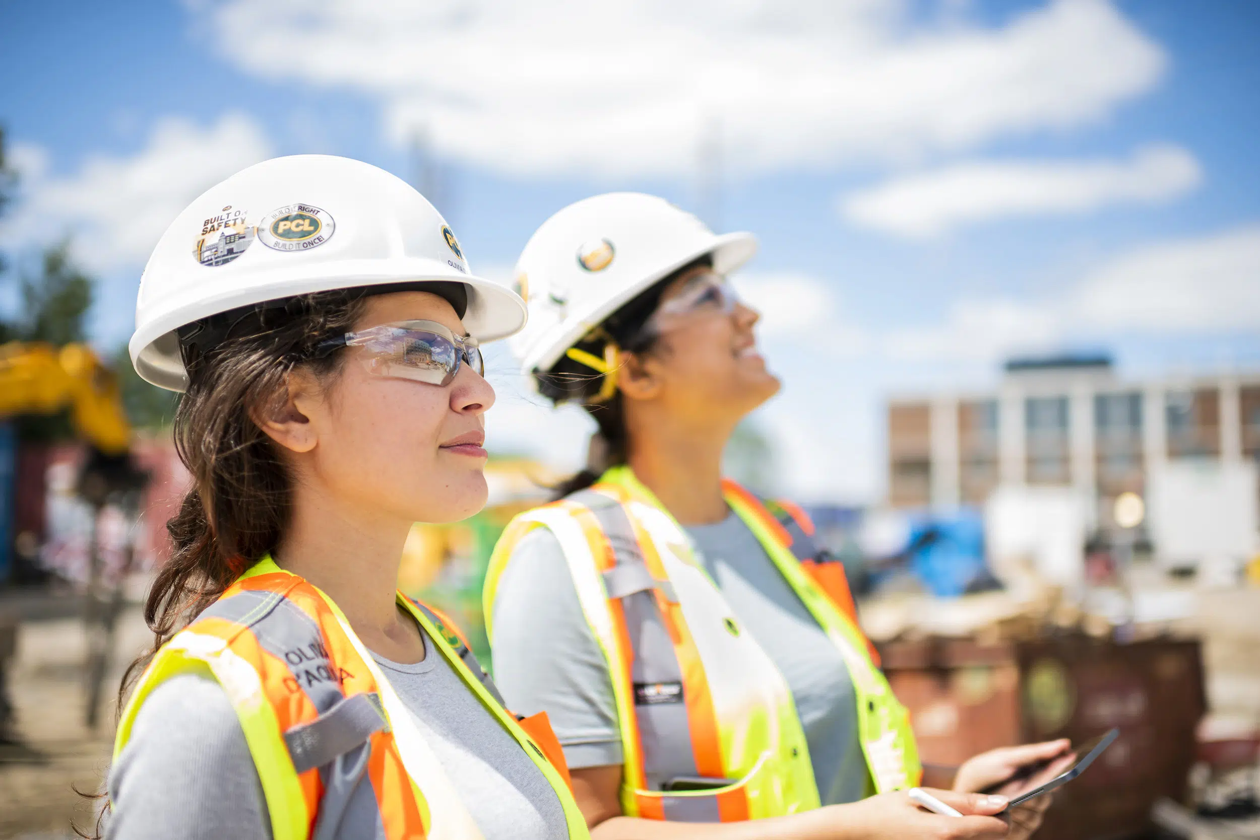 Two young female construction workers are pictured looking in the distance. One holds a tablet.