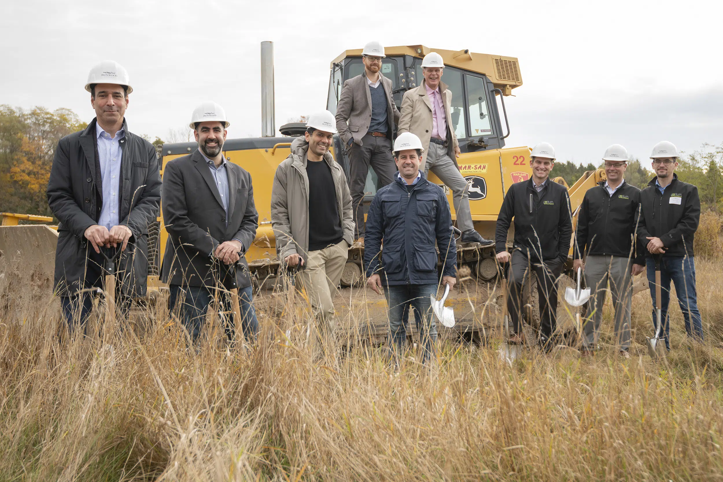 A team of developers pose for a group photo on a bulldozer. They are seen standing in long grass holding shovels and wearing hard hats.